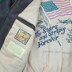 Puede incluir: Una chaqueta de cuero marr&oacute;n oscuro con forro interior color crema. El forro presenta un gr&aacute;fico de la bandera estadounidense, aviones, el texto The Stars and Stripes Forever, Avirex Ltd y un parche rectangular con la etiqueta Italy 1 Lira.