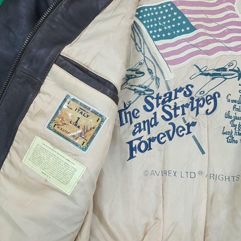 Puede incluir: Una chaqueta de cuero marr&oacute;n oscuro con forro interior color crema. El forro presenta un gr&aacute;fico de la bandera estadounidense, aviones, el texto The Stars and Stripes Forever, Avirex Ltd y un parche rectangular con la etiqueta Italy 1 Lira.