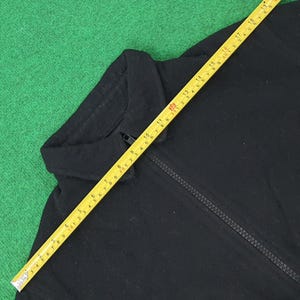 K&ouml;nnte beinhalten: Schwarze Jacke mit Rei&szlig;verschluss und Kragen. Ein gelbes Ma&szlig;band liegt &uuml;ber der Jacke und zeigt Ma&szlig;e in Zentimetern an. Die Jacke liegt auf einer gr&uuml;nen Oberfl&auml;che.