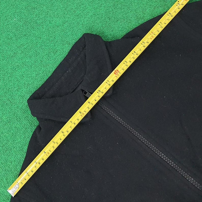 K&ouml;nnte beinhalten: Schwarze Jacke mit Rei&szlig;verschluss und Kragen. Ein gelbes Ma&szlig;band liegt &uuml;ber der Jacke und zeigt Ma&szlig;e in Zentimetern an. Die Jacke liegt auf einer gr&uuml;nen Oberfl&auml;che.