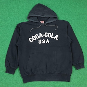 Può includere: Felpa con cappuccio nera con il logo Coca-Cola bianco sul petto. La felpa ha una tasca frontale e un cappuccio con coulisse. Il testo "USA" è sotto il logo Coca-Cola.