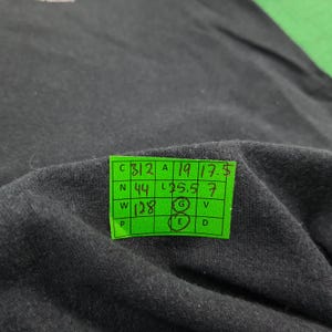 May include: A black fabric garment featuring a bright green rectangular measurement tag with a grid layout. The tag displays handwritten numbers including 312, 19, 17.5, 44, 25.5, 7, and 128, with the letters G and E circled in black ink on the grid.