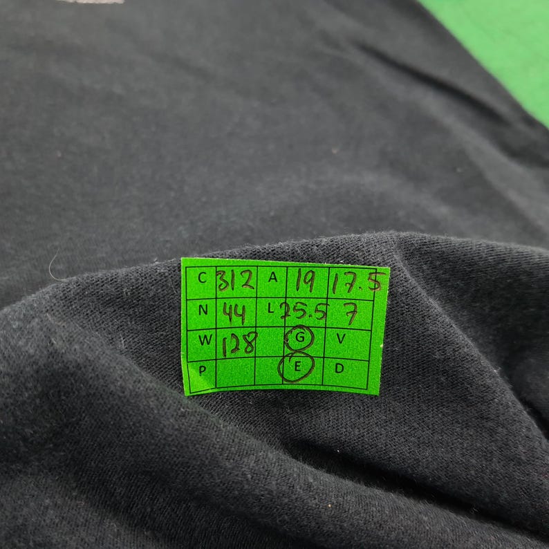 May include: A black fabric garment featuring a bright green rectangular measurement tag with a grid layout. The tag displays handwritten numbers including 312, 19, 17.5, 44, 25.5, 7, and 128, with the letters G and E circled in black ink on the grid.