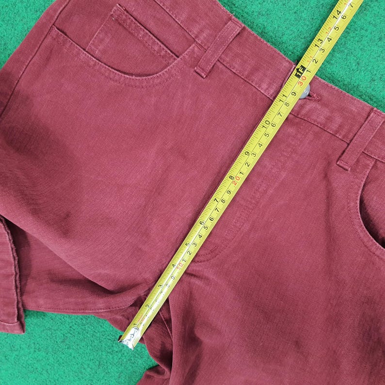 May include: Close-up of burgundy-colored denim pants with a yellow measuring tape. The tape shows a measurement of approximately 14 inches. The pants have a classic five-pocket design and are laid on a green surface.