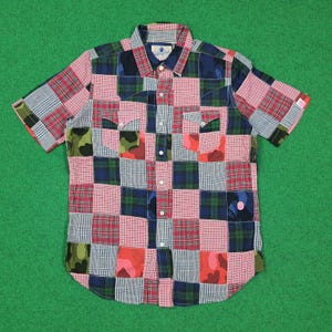 May include: A short-sleeve button-down shirt featuring a patchwork design composed of various plaid, checkered, and camouflage patterns in shades of red, blue, green, and white. The shirt includes two chest pockets with flaps and white buttons.