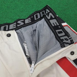 May include: Close-up of motorcycle pants in cream color with a red stripe and black accents. The pants have a zipper, snap closure, and a mesh lining. The waistband features the brand name in gray and black.