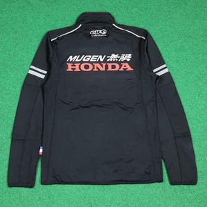May include: Black zip-up jacket with white and red text that reads "MUGEN HONDA" and Japanese characters. The jacket has white stripes on the sleeves and the "elf Lubrifiants" logo. The jacket is on a green surface.
