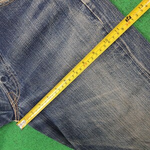 K&ouml;nnte beinhalten: Nahaufnahme einer blauen Jeans mit einem gelben Ma&szlig;band. Das Band zeigt Ma&szlig;e in Zentimetern, wobei die Stoffstruktur und die Verarbeitung des Kleidungsst&uuml;cks hervorgehoben werden. Die Jeans liegt auf einer gr&uuml;nen Oberfl&auml;che.