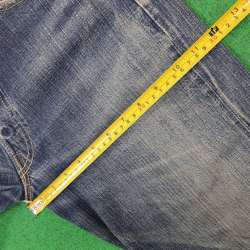 K&ouml;nnte beinhalten: Nahaufnahme einer blauen Jeans mit einem gelben Ma&szlig;band. Das Band zeigt Ma&szlig;e in Zentimetern, wobei die Stoffstruktur und die Verarbeitung des Kleidungsst&uuml;cks hervorgehoben werden. Die Jeans liegt auf einer gr&uuml;nen Oberfl&auml;che.
