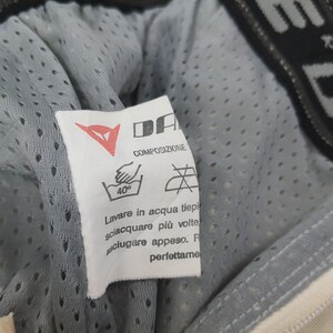 May include: A close-up of a white tag with the brand logo "DA" and washing instructions. The tag is attached to a gray, perforated fabric. The text on the tag includes "COMPOSIZIONE" and washing symbols.