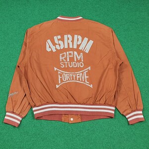 May include: An orange bomber jacket with white and red striped trim on the cuffs and waist. The back of the jacket features the text "45RPM STUDIO FORTY FIVE" in a distressed white font. The jacket has a button closure and a white collar.
