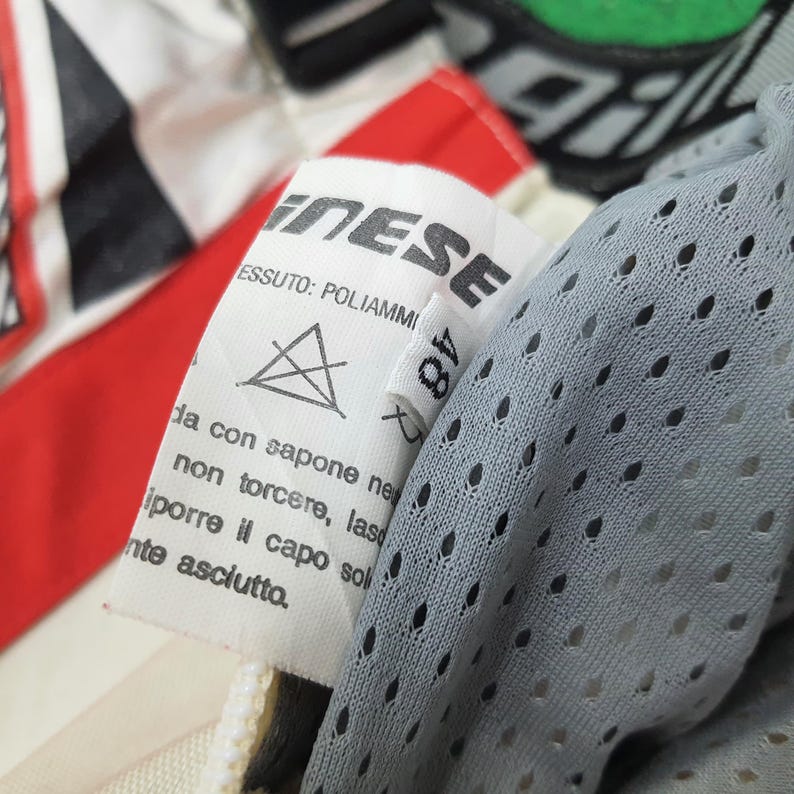 May include: Close-up of a white label with the brand name "NESE" and the number "48". The label is attached to a garment with red, white, and black accents. A gray, perforated fabric is visible on the right side.