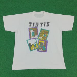 May include: White t-shirt featuring the text "TINTIN" above four colorful rectangular illustrations. The illustrations depict scenes with characters and dogs. The shirt is laid flat on a green surface.