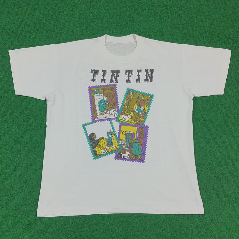 May include: White t-shirt featuring the text "TINTIN" above four colorful rectangular illustrations. The illustrations depict scenes with characters and dogs. The shirt is laid flat on a green surface.