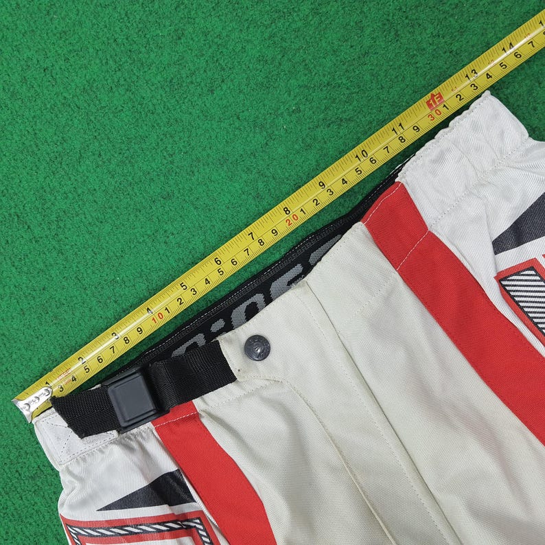 May include: Close-up of motorcycle pants in cream, red, and black. The pants have a black belt with a buckle and a button closure. A yellow measuring tape is laid across the pants, showing measurements in inches.
