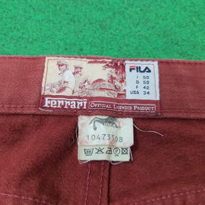May include: Close-up of a pair of burgundy pants with a FILA and Ferrari label. The labels include text and graphics, with size information and the words "OFFICIAL LICENSED PRODUCT". The pants are on a green surface.