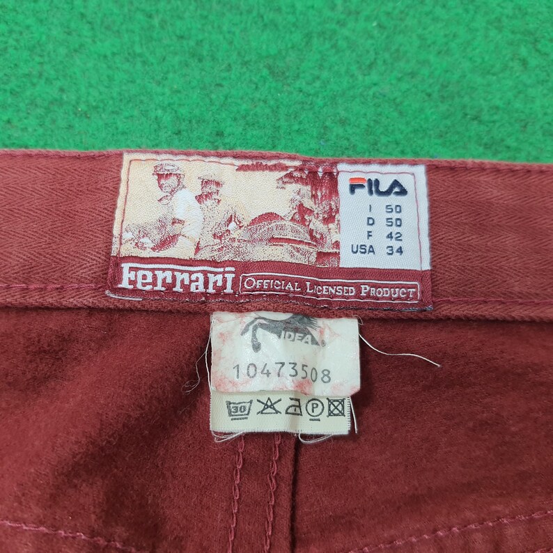 May include: Close-up of a pair of burgundy pants with a FILA and Ferrari label. The labels include text and graphics, with size information and the words "OFFICIAL LICENSED PRODUCT". The pants are on a green surface.