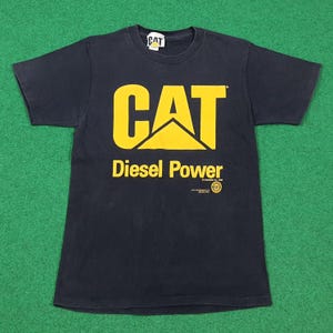 May include: A black short-sleeve t-shirt featuring the yellow CAT logo and the text Diesel Power printed on the front. The shirt includes a small circular logo and copyright text below the main graphic, with a CAT brand tag at the inner neckline.