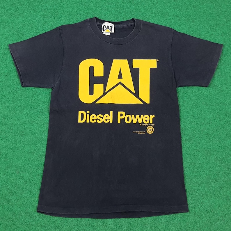 May include: A black short-sleeve t-shirt featuring the yellow CAT logo and the text Diesel Power printed on the front. The shirt includes a small circular logo and copyright text below the main graphic, with a CAT brand tag at the inner neckline.