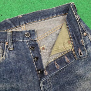 K&ouml;nnte beinhalten: Nahaufnahme einer blauen Jeans mit Knopfleiste. Die Jeans hat schwarze Kn&ouml;pfe und sichtbare N&auml;hte in einem kontrastierenden gelben Faden. Der Hosenschlitz ist teilweise aufgekn&ouml;pft und gibt den Blick auf den Innenstoff frei.