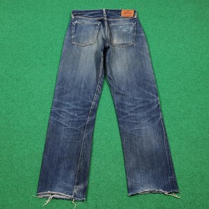 K&ouml;nnte beinhalten: Verblasste blaue Jeans aus Denim mit einem Used-Look, die sichtbare Abnutzungsmuster und einen rohen Saum aufweisen. Die Jeans haben ein klassisches F&uuml;nf-Taschen-Design und einen Lederaufn&auml;her am hinteren Bund. Die Jeans werden auf einer gr&uuml;nen Oberfl&auml;che pr&auml;sentiert.