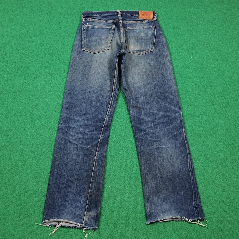 K&ouml;nnte beinhalten: Verblasste blaue Jeans aus Denim mit einem Used-Look, die sichtbare Abnutzungsmuster und einen rohen Saum aufweisen. Die Jeans haben ein klassisches F&uuml;nf-Taschen-Design und einen Lederaufn&auml;her am hinteren Bund. Die Jeans werden auf einer gr&uuml;nen Oberfl&auml;che pr&auml;sentiert.