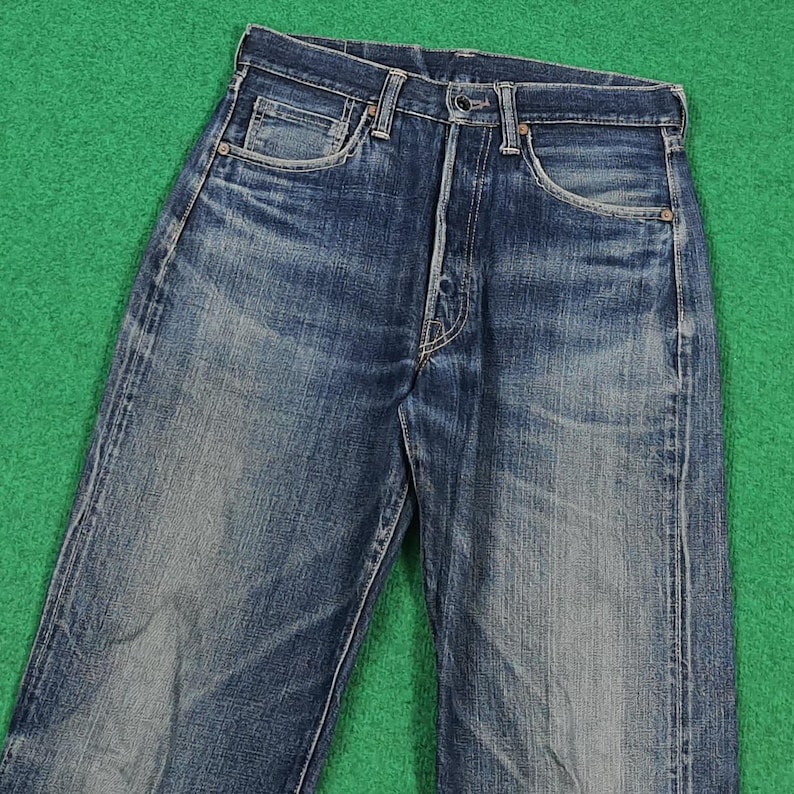 K&ouml;nnte beinhalten: Verblasste blaue Jeans mit Knopfleiste, F&uuml;nf-Taschen-Design und klassischer gerader Beinpassform. Die Jeans weisen Gebrauchsspuren und Ausbleichungen auf, was ihnen einen Vintage-Look verleiht. Der Hintergrund ist einfarbig gr&uuml;n.