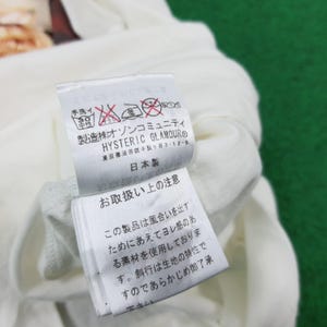 Puede incluir: Una etiqueta de prenda de tela blanca con texto en japon&eacute;s negro y el nombre de la marca HYSTERIC GLAMOUR. La etiqueta incluye varios s&iacute;mbolos de cuidado de lavado, incluido un icono de lavado a mano, e indica que el producto es hecho en Jap&oacute;n.