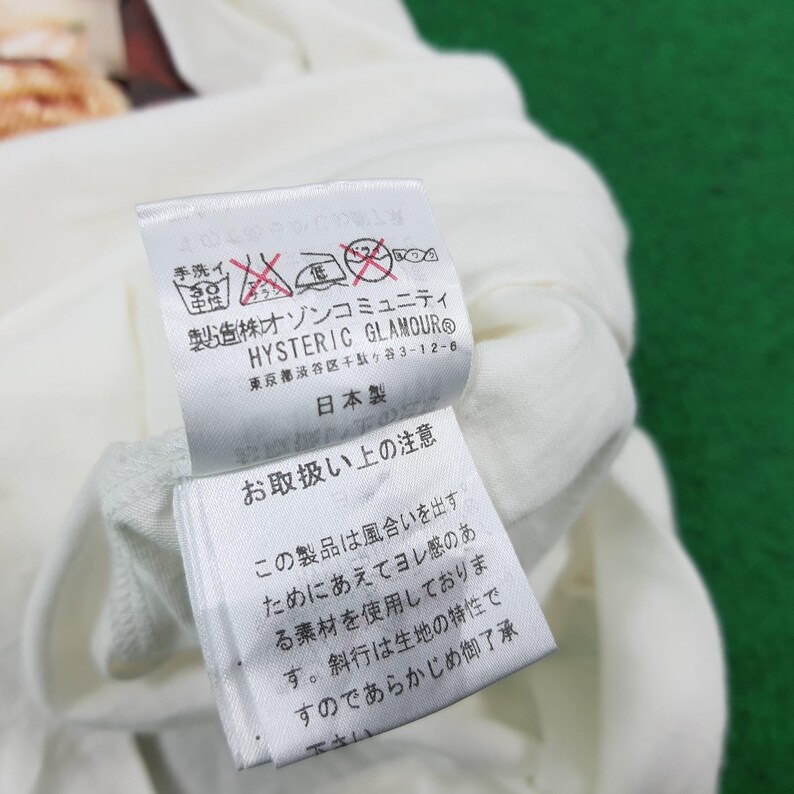 Puede incluir: Una etiqueta de prenda de tela blanca con texto en japon&eacute;s negro y el nombre de la marca HYSTERIC GLAMOUR. La etiqueta incluye varios s&iacute;mbolos de cuidado de lavado, incluido un icono de lavado a mano, e indica que el producto es hecho en Jap&oacute;n.