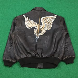 Puede incluir: Una chaqueta bomber de cuero marr&oacute;n oscuro con un gran gr&aacute;fico pintado a mano en la espalda. El dise&ntilde;o muestra una mano alada de color beige y blanco que sostiene una pluma estilogr&aacute;fica con una punta plateada. La chaqueta tiene pu&ntilde;os y dobladillo negros.