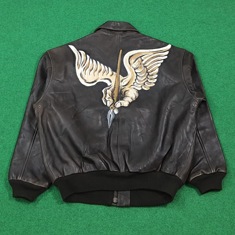 Puede incluir: Una chaqueta bomber de cuero marr&oacute;n oscuro con un gran gr&aacute;fico pintado a mano en la espalda. El dise&ntilde;o muestra una mano alada de color beige y blanco que sostiene una pluma estilogr&aacute;fica con una punta plateada. La chaqueta tiene pu&ntilde;os y dobladillo negros.