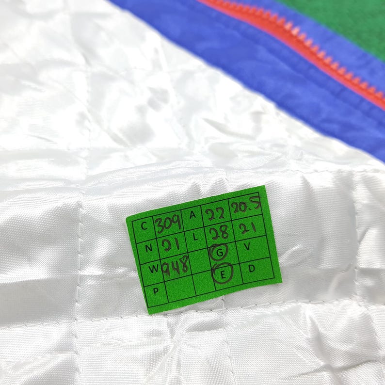May include: Close-up of a white quilted fabric with a green tag. The tag has black text and numbers, including "309", "A 22 20.5", "N 21", "L 28 21", and "W 948". The fabric has a subtle sheen.