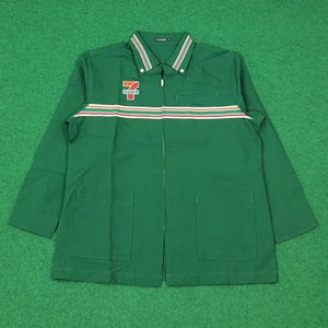 Puede incluir: Chaqueta verde con cremallera y el logotipo de 7-Eleven en el pecho. La chaqueta tiene un cuello blanco con rayas verdes y un diseño de rayas horizontales en rojo, blanco y naranja en el pecho. Se ven dos bolsillos grandes.
