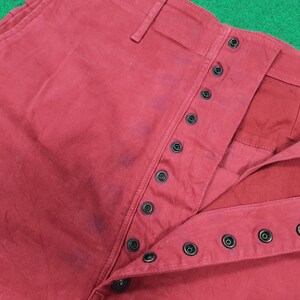 May include: Close-up of red pants with a button-up fly. The pants have a classic design with multiple black buttons and a visible waistband. The fabric appears to be a sturdy cotton or similar material.