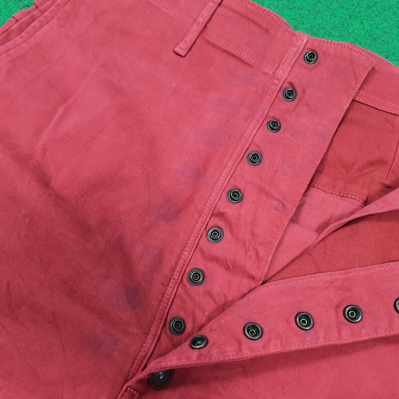May include: Close-up of red pants with a button-up fly. The pants have a classic design with multiple black buttons and a visible waistband. The fabric appears to be a sturdy cotton or similar material.