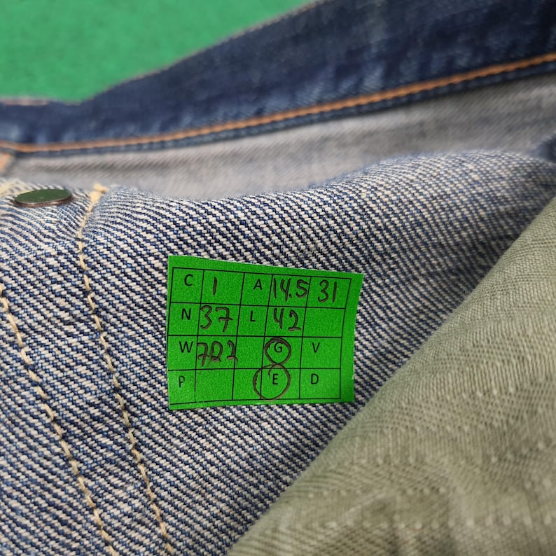 K&ouml;nnte beinhalten: Nahaufnahme von Jeans aus Denim mit einem gr&uuml;nen Etikett. Das Etikett enth&auml;lt handschriftliche Ma&szlig;e und Zahlen. Die Jeans ist blau mit sichtbaren N&auml;hten und einem Knopf. Der Stoff ist ein mittelblaues Denim.