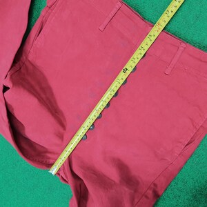May include: Close-up of red pants with a row of black buttons down the side. A yellow measuring tape is laid across the pants, showing measurements in inches. The pants are made of a solid, matte fabric.