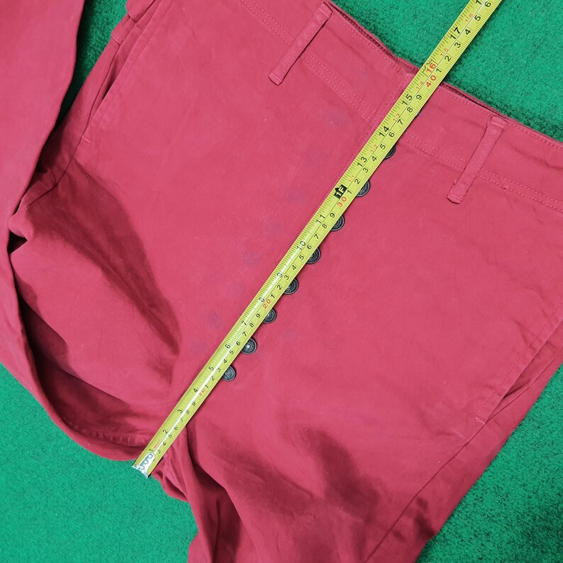 May include: Close-up of red pants with a row of black buttons down the side. A yellow measuring tape is laid across the pants, showing measurements in inches. The pants are made of a solid, matte fabric.