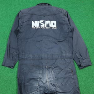 May include: Dark blue jumpsuit with the white text "NISMO NISSAN RACING TEAM" on the back. The jumpsuit has long sleeves, a button-down front, and multiple pockets. The overall design is a workwear style.