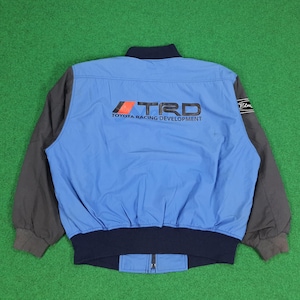 Puede incluir: Una chaqueta azul claro con mangas gris oscuro y cuello y dobladillo acanalados en azul marino. La espalda presenta el texto negro TRD Toyota Racing Development con un elemento gráfico rojo y negro. Un pequeño parche negro está en la manga.
