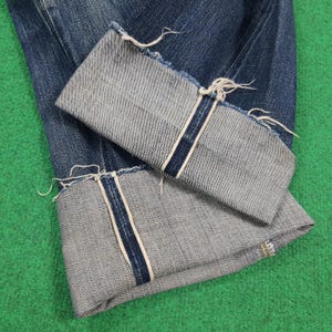 K&ouml;nnte beinhalten: Nahaufnahme einer blauen Jeans mit hochgekrempelten Manschetten. Die Manschetten zeigen eine hellere Denim-Innenseite mit einem marineblauen Streifen und ausgefransten Kanten. Die Jeans liegt auf einer gr&uuml;nen Oberfl&auml;che.