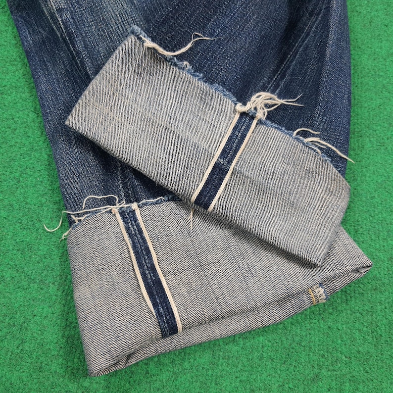 K&ouml;nnte beinhalten: Nahaufnahme einer blauen Jeans mit hochgekrempelten Manschetten. Die Manschetten zeigen eine hellere Denim-Innenseite mit einem marineblauen Streifen und ausgefransten Kanten. Die Jeans liegt auf einer gr&uuml;nen Oberfl&auml;che.
