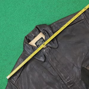 Puede incluir: Una chaqueta de vuelo de cuero marr&oacute;n oscuro tipo A-2 con una etiqueta interior beige que dice AVIREX. Una cinta m&eacute;trica amarilla est&aacute; colocada diagonalmente sobre la chaqueta, mostrando una longitud de aproximadamente 63,5 cm desde el cuello al dobladillo.