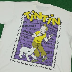 May include: White t-shirt featuring a graphic of Tintin, a cartoon character, and his dog Snowy. The design includes the word "TINTIN" in yellow, a purple stamp-like background, and newspaper-style text.