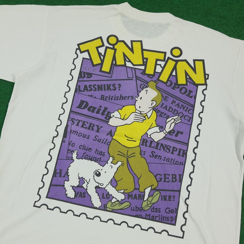 May include: White t-shirt featuring a graphic of Tintin, a cartoon character, and his dog Snowy. The design includes the word "TINTIN" in yellow, a purple stamp-like background, and newspaper-style text.