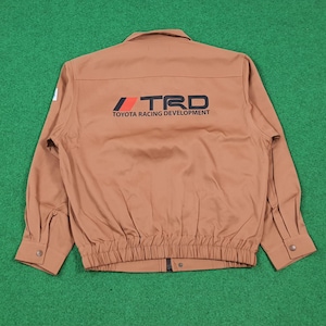 May include: A tan long-sleeve work jacket featuring a collar, buttoned cuffs, and an elasticized hem. The back displays the black and red TRD Toyota Racing Development logo printed in the center. The jacket has a front zipper closure.