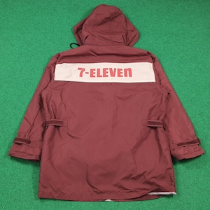 Puede incluir: Chaqueta con capucha de color granate con el texto "7-ELEVEN" en letras rojas sobre un parche rectangular blanco en la parte superior de la espalda. La chaqueta tiene capucha, dos bolsillos y puños ajustables. El fondo es verde.