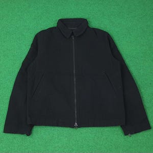 K&ouml;nnte beinhalten: Schwarze Jacke mit Kragen und durchgehendem Rei&szlig;verschluss. Die Jacke hat zwei Vordertaschen und lange &Auml;rmel. Der Hintergrund ist einfarbig gr&uuml;n.