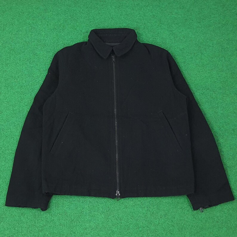 K&ouml;nnte beinhalten: Schwarze Jacke mit Kragen und durchgehendem Rei&szlig;verschluss. Die Jacke hat zwei Vordertaschen und lange &Auml;rmel. Der Hintergrund ist einfarbig gr&uuml;n.