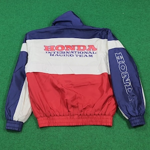Può includere: Una giacca Honda International Racing Team blu, bianca e rossa. La giacca ha un cappuccio e un orlo a coste. Il logo Honda è stampato sul petto e sulla manica.
